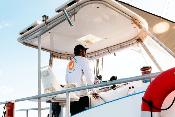 Person steering a boat from a covered cockpit with a lifebuoy and clear sky.