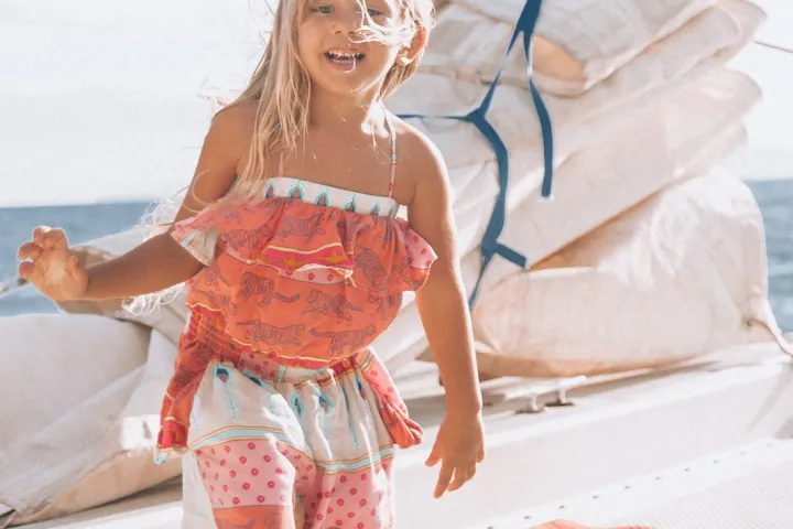 Smiling child in colorful outfit on a boat with sails and ocean in background.