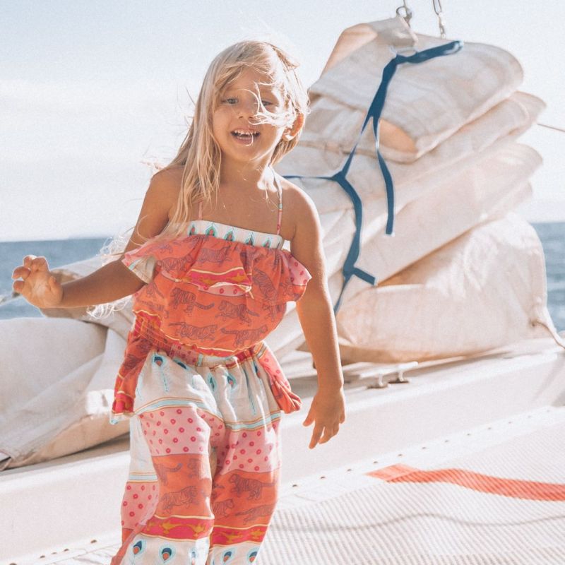 Smiling child in colorful outfit on a boat with sails and ocean in background.
