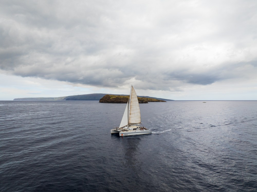 Sailboat on calm sea with distant islands under a cloudy sky.