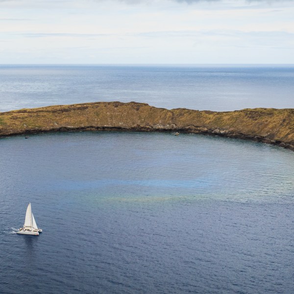 Aerial view of a crescent-shaped island with a lone sailboat nearby on the ocean.