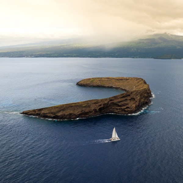 Aerial view of a sailboat near a crescent-shaped island in a vast ocean.