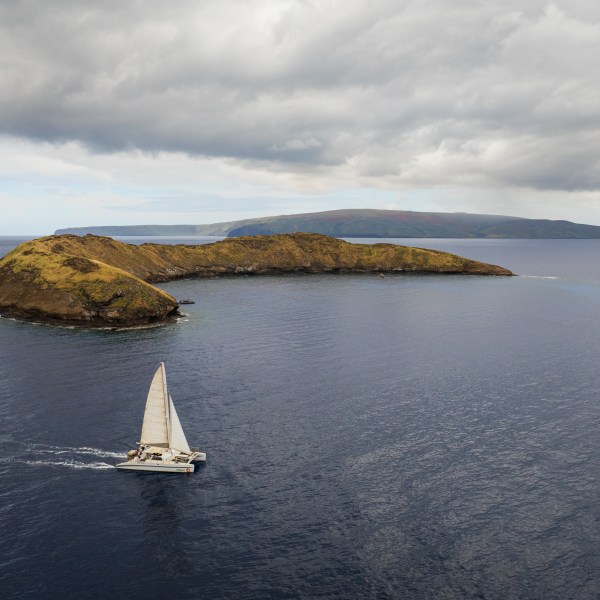 A sailboat near a green island under cloudy skies, surrounded by vast ocean.