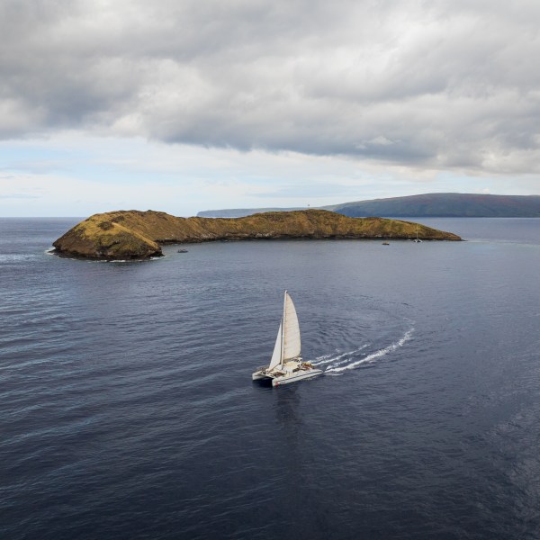 Sailboat on ocean with a small island under cloudy sky.