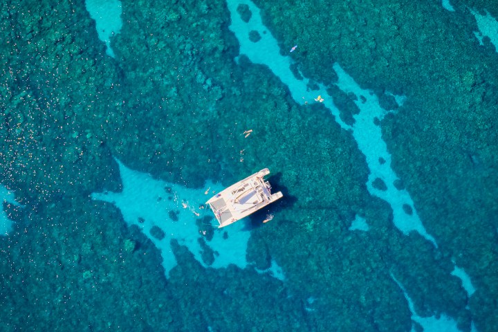Aerial view of a boat on turquoise ocean over a coral reef with visible swimmers.