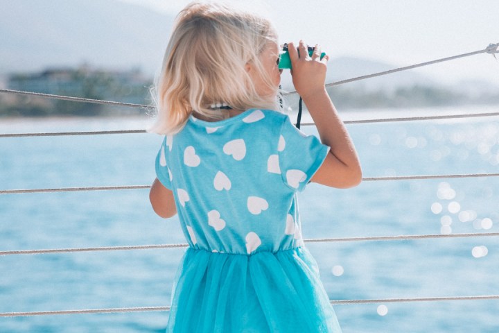 Child in blue dress looking through toy binoculars over water.