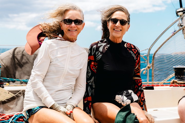 Two women smiling on a boat, wearing sunglasses and outdoor clothing, with a sunny sky in the background.