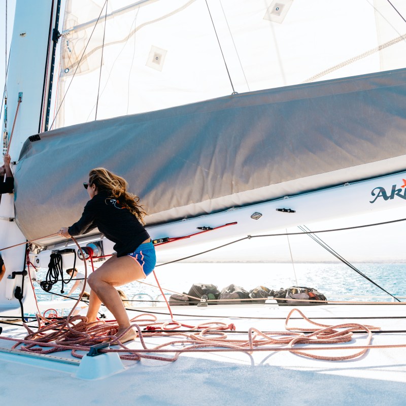 Two people on a boat adjusting ropes near a sail with 'Akaloga' written on it.