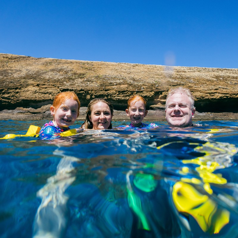 Family snorkeling in clear water near a rocky shore.