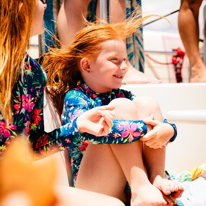 Two red-haired kids in floral swimsuits sitting on a boat, smiling.