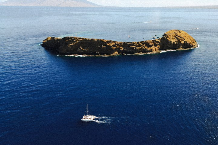 Aerial view of a catamaran near a small island with blue ocean and distant mountains.