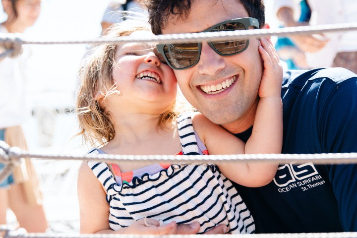 Man with sunglasses hugging a smiling child in striped dress on a boat.