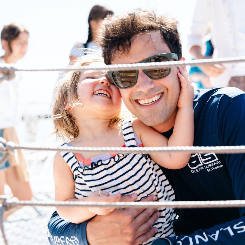 Man with sunglasses hugging a smiling child in striped dress on a boat.