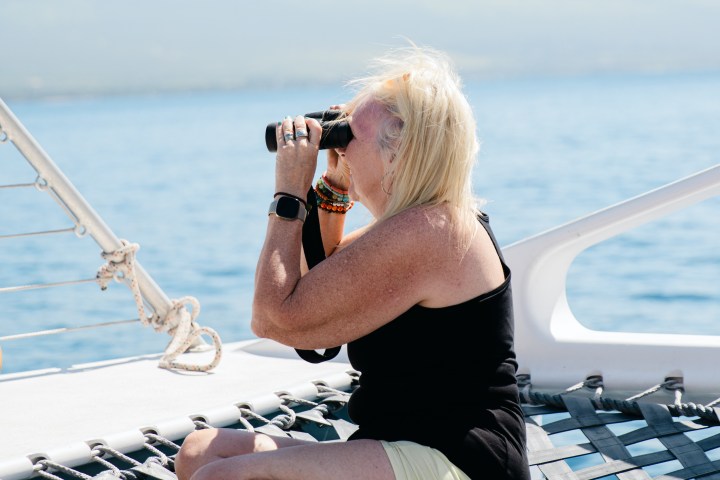 Person on boat using binoculars, sitting on netting with ocean and mountains in background.