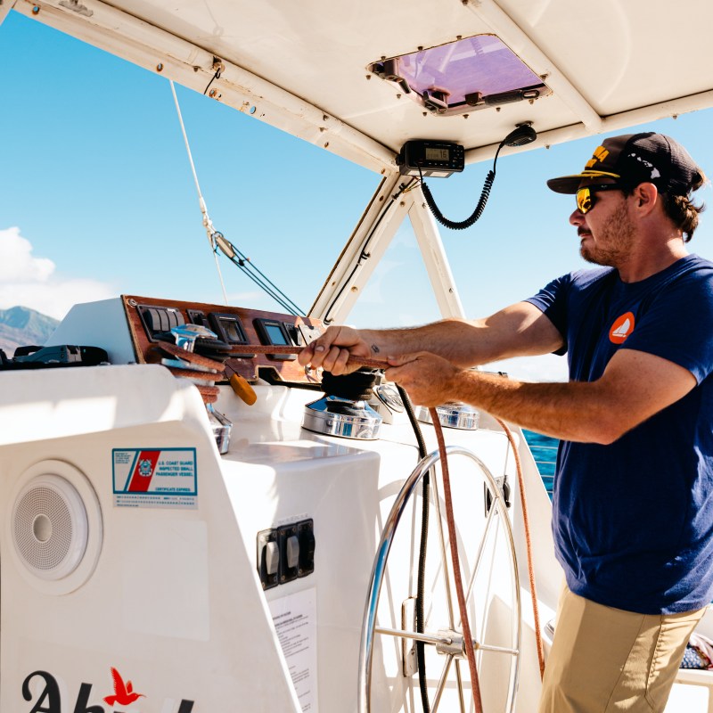 Man in sunglasses and cap steering a boat with visible control panel and wheel.