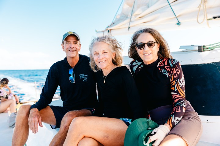 Three adults sitting on a boat, smiling and wearing sun protection clothing under a clear blue sky.