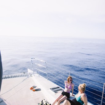 Two people sitting on a boat deck, overlooking calm blue ocean under a clear sky.