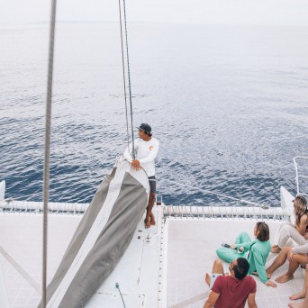 People sitting on a catamaran's deck with one person adjusting sails on calm water.