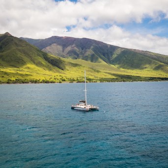 Catamaran on blue ocean with green mountainous background under cloudy sky.
