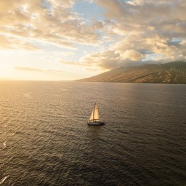 Sailboat on open sea at sunset with mountains and clouds in the background.