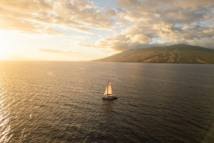 Sailboat on open sea at sunset with mountains and clouds in the background.