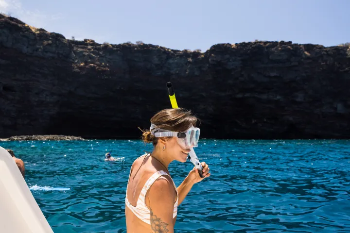 Woman in swimwear holding snorkel sitting on boat deck with flippers near blue ocean and cliffs.