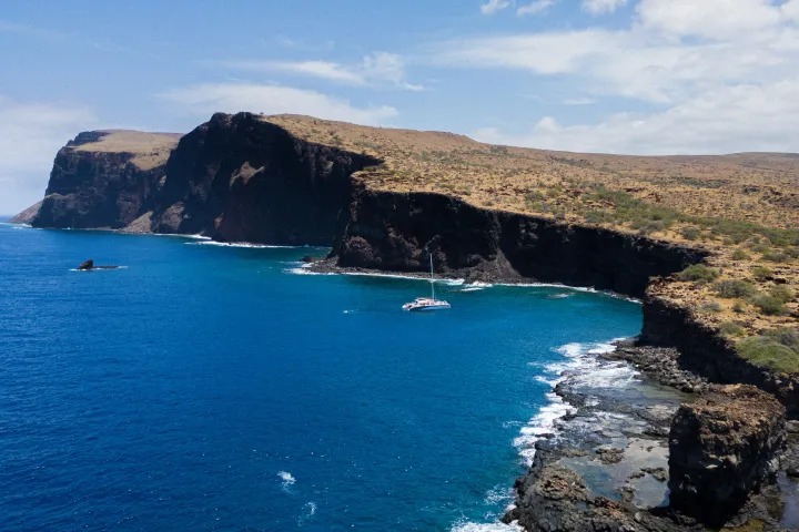 Aerial view of a sailboat near rocky cliffs and the open sea under a partly cloudy sky.