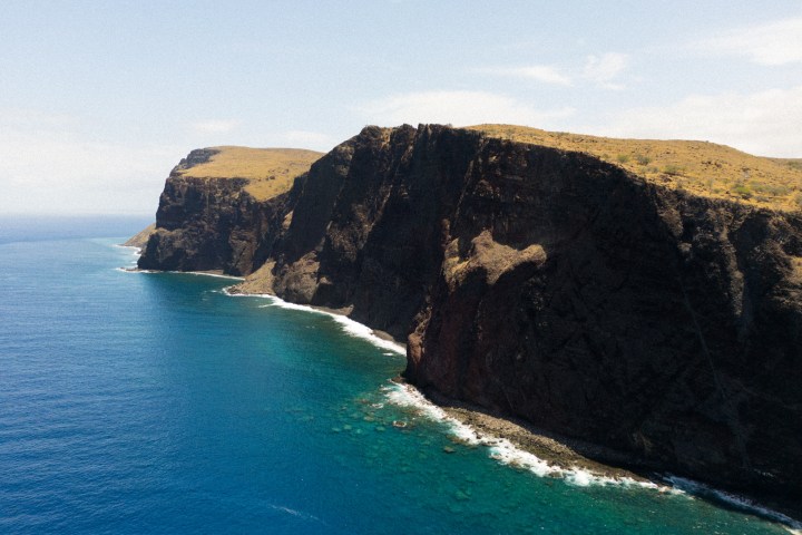 Cliffside view of ocean with rugged terrain and blue water under a partly cloudy sky.