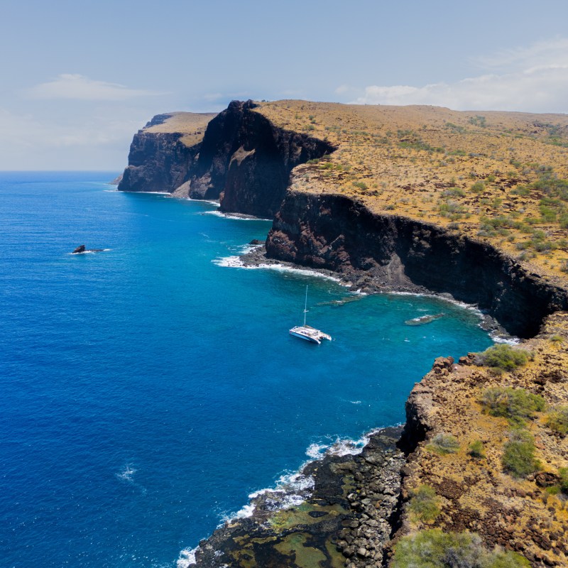 Aerial view of a sailboat near a steep, rocky coastline with clear blue water and a partly cloudy sky.