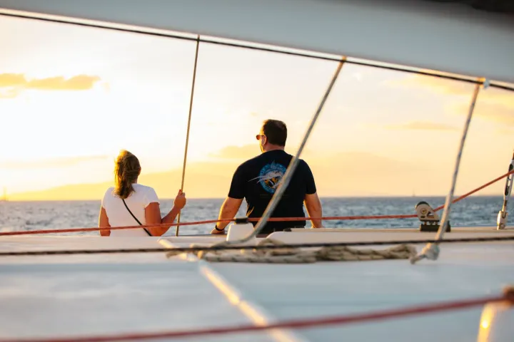 Two people sitting on a sailboat deck at sunset, facing the ocean.