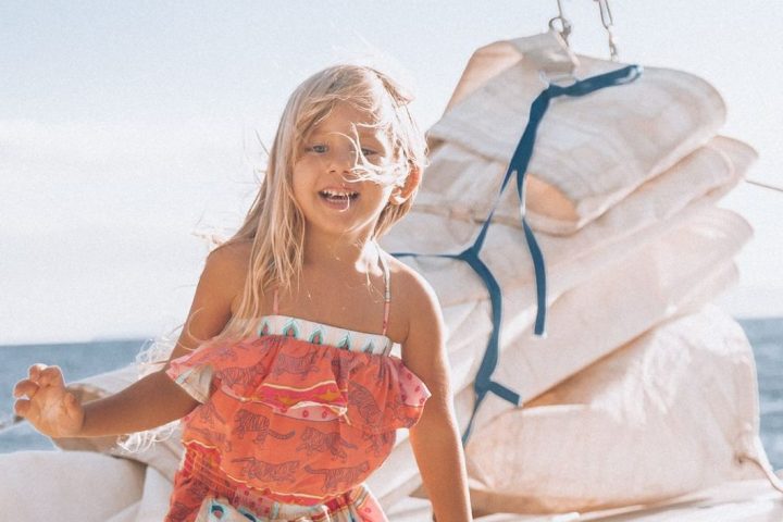 Smiling child on a sailboat deck wearing colorful clothing, with sails in the background.