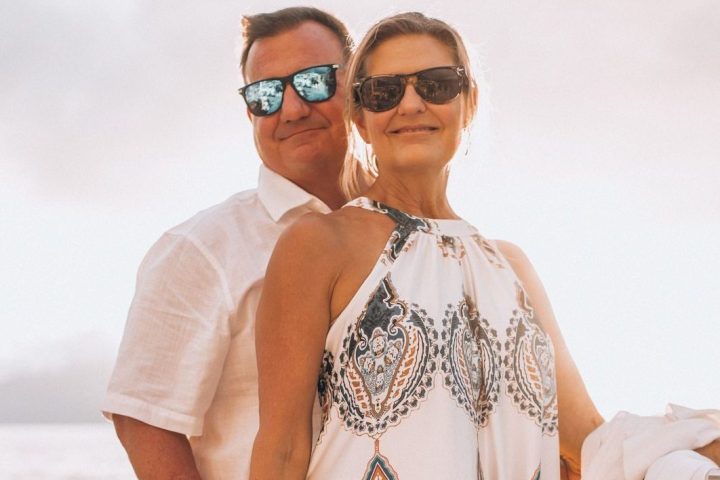 Couple in sunglasses on a boat, woman in a patterned dress, ocean in background.
