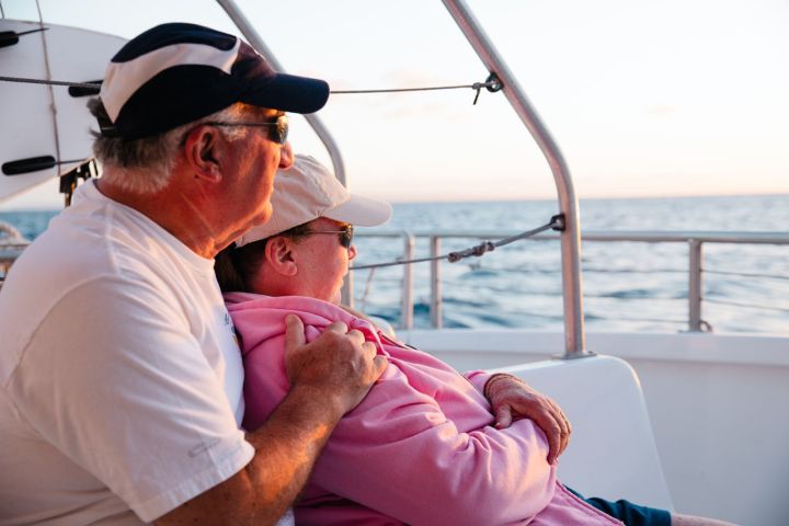 Elderly couple in hats embraces while sitting on a boat, gazing at the ocean during sunset.