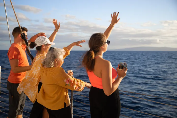 Group of people waving from a boat, ocean and distant land in background, sunny day.