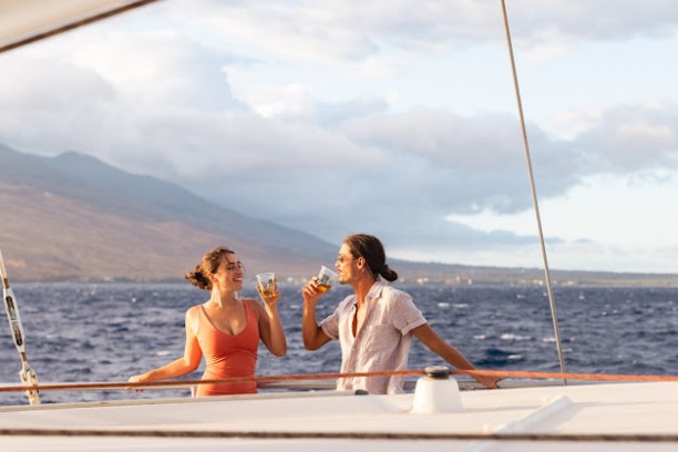 Two people on a boat enjoy drinks with a mountainous backdrop and ocean view.