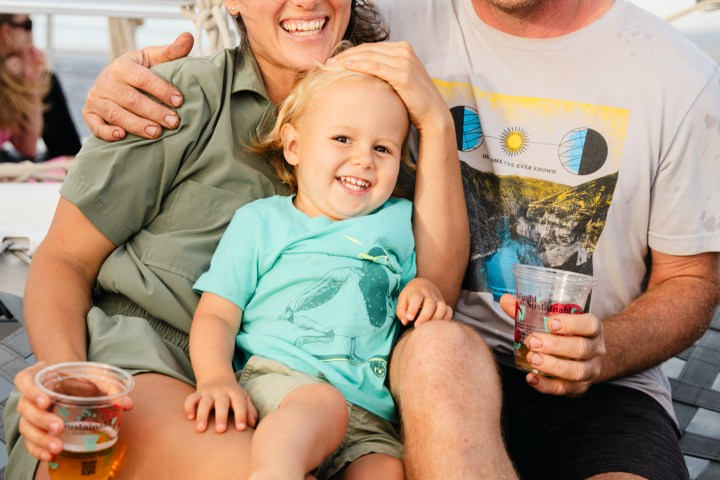 Smiling family on a boat, woman, man, and child holding drinks, sunny day.