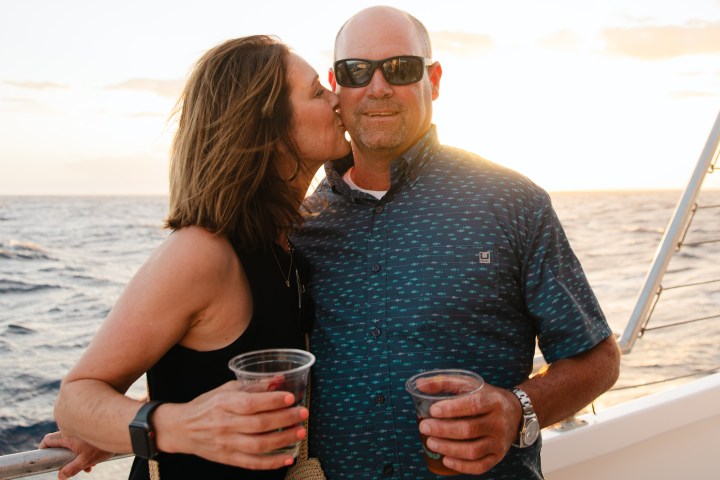 Two people on a boat at sunset, one kissing the other's cheek, holding drinks.