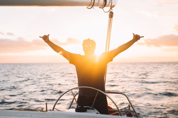 Person on boat at sunset with arms raised, sun behind.