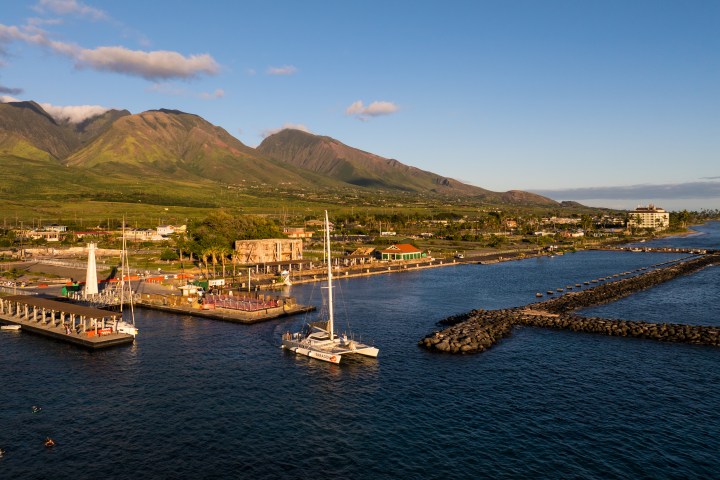 Coastal town with docks, sailboat, mountains, and clear sky at sunset.